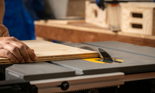 Person using a table saw with a piece of wood on a workshop table.