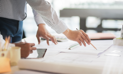An individual with pencil in hand pointing at blueprints spread across a table